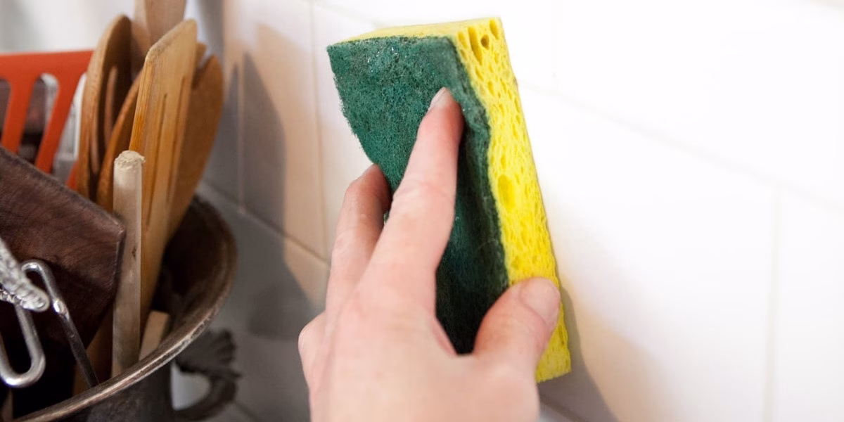 A hand holding a sponge cleaning a greasy kitchen backsplash